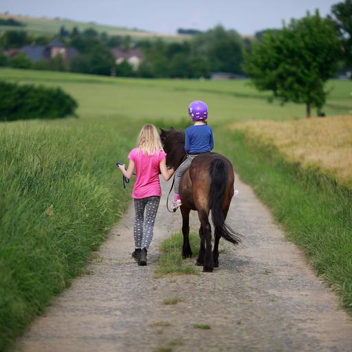 Ein Maedchen reitet unter Aufsicht mit einem Pony auf einem Feldweg aus.