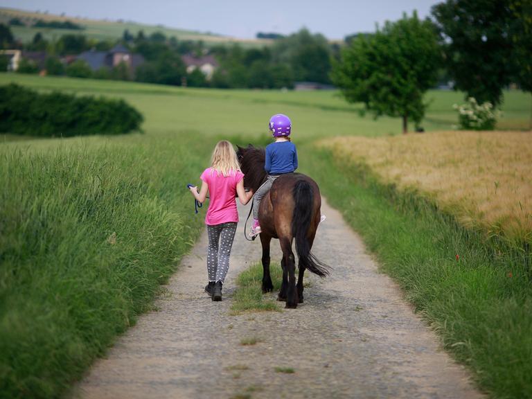 Ein Maedchen reitet unter Aufsicht mit einem Pony auf einem Feldweg aus.