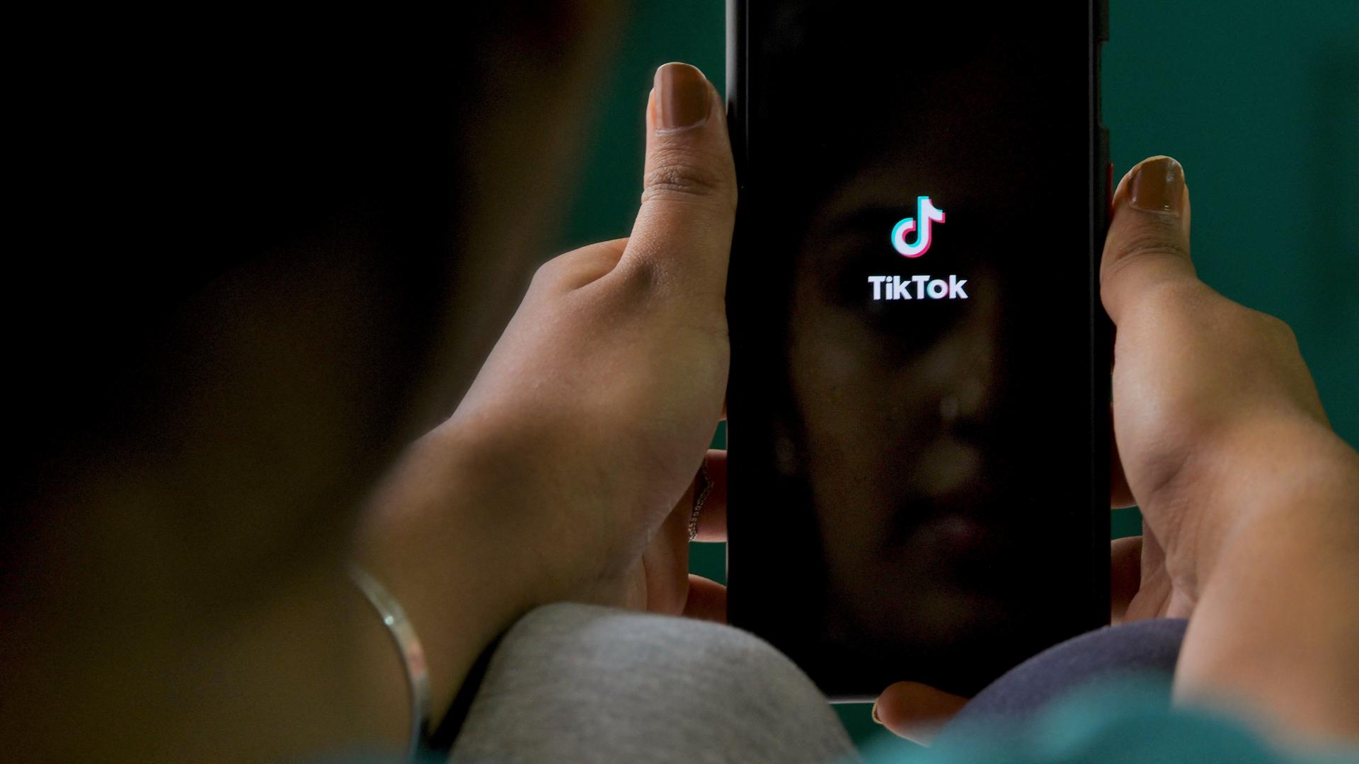 Nahaufnahme von Händen, die ein Smartphone mit geöffneter Tiktok-App halten.