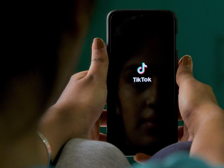 Nahaufnahme von Händen, die ein Smartphone mit geöffneter Tiktok-App halten.