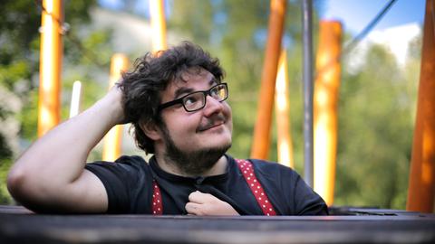 Ein junger Mann mit dunklem, gelocktem Haar und schwarz umrandeter Brille stützt den Kopf auf den Ellbogen. Im Hintergrund sind Bäume und Stämme zu sehen. Er trägt ein schwarzes T-Shirt und rote Hosenträger mit weißen Punkten. Sein Blick ist verträumt lächelnd zur Seite und nach oben gerichtet.