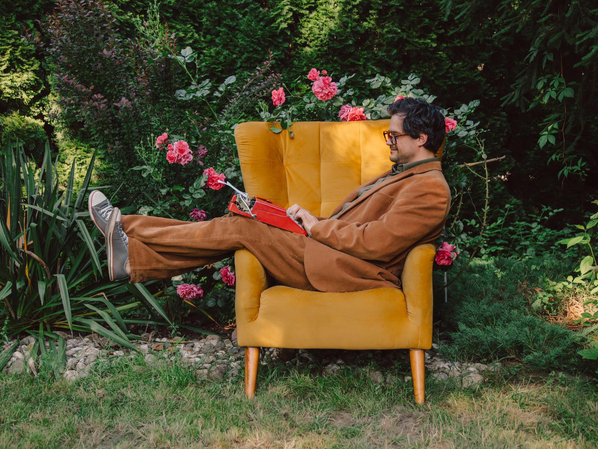 Ein Mann im Anzug liegt im Garten auf einem Sessel und tippt auf einer Schreibmaschine