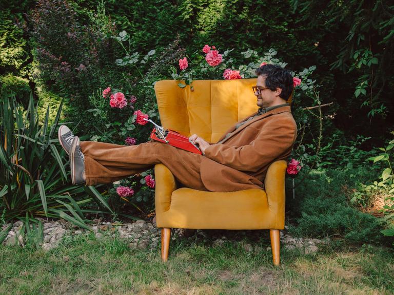 Ein Mann im Anzug liegt im Garten auf einem Sessel und tippt auf einer Schreibmaschine