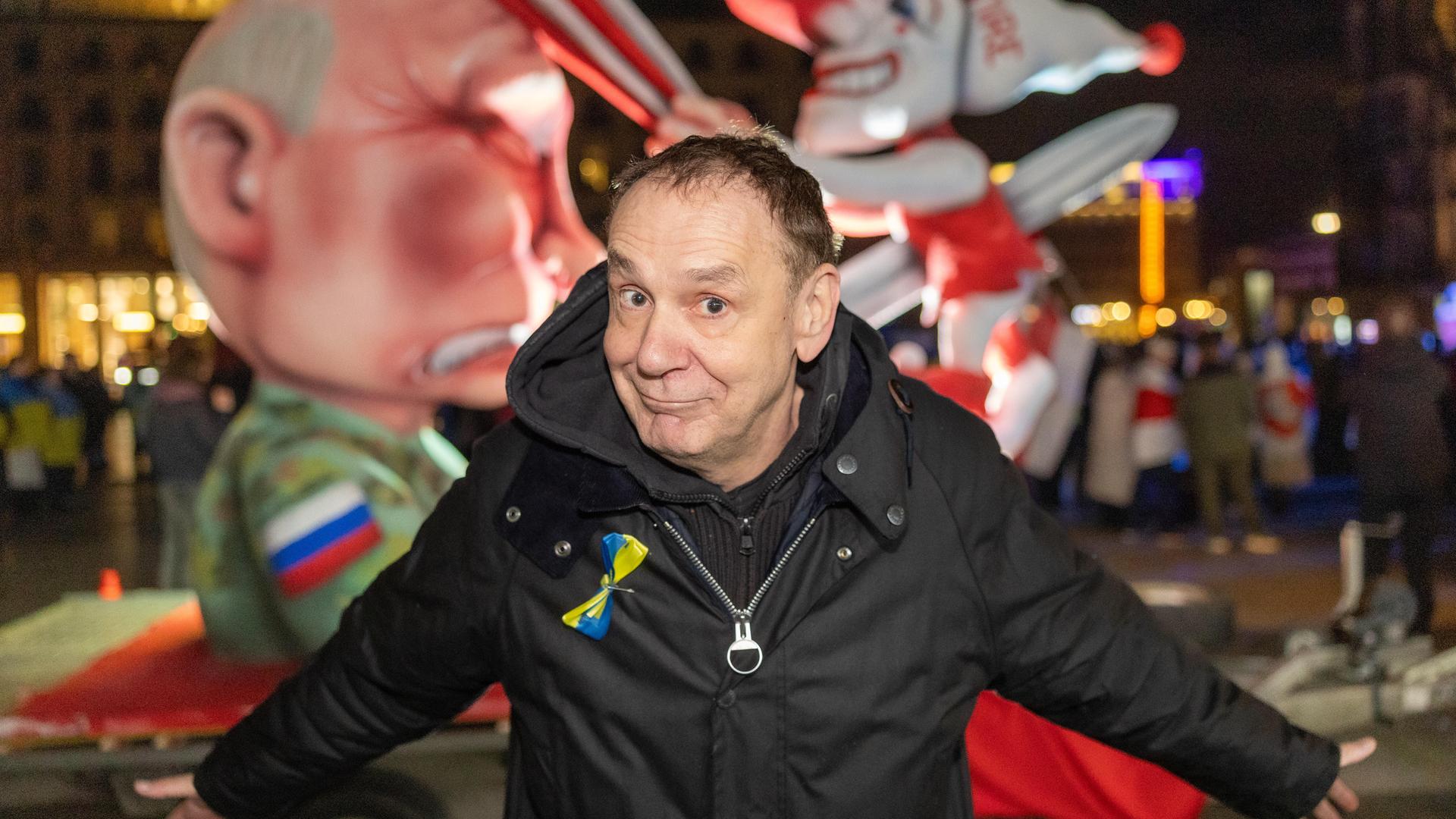 Tilly steht vor dem Wagen und breitet mit spöttischem Lächeln beide Arme aus. Es ist dunkel. Dahinter der Wagen, in dem eine Putin-Figur den Düsseldorfer Schalk "Hoppeditz" mit einem Schwert aufspießt. Dieser schlägt Putin mit einer Klatsche auf den Kopf.