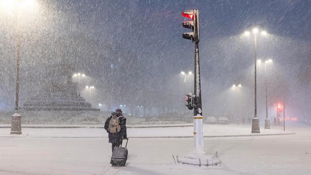 Ein Mann zieht einen Roll-Koffer durch den Schnee in Paris. Es sind keine Autos auf den Straßen. Eine Ampel leuchtet rot.