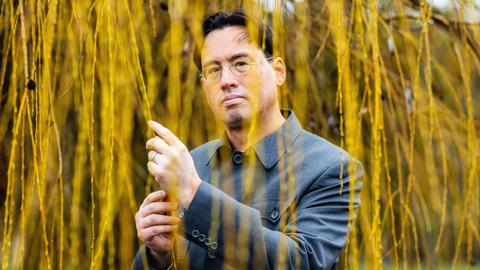 Ein Mann mit chinesisch-amerikanischen Wurzeln steht unter einem Baum und hält herabhängende Weidenzweige in der Hand Ein Mann mit chinesisch-amerikanischen Wurzeln steht unter einem Baum und hält herabhängende Weidenzweige in der Hand