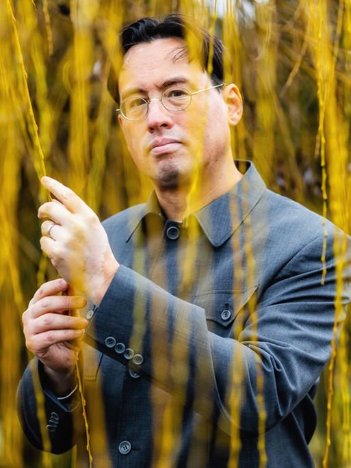 Ein Mann mit chinesisch-amerikanischen Wurzeln steht unter einem Baum und hält herabhängende Weidenzweige in der Hand Ein Mann mit chinesisch-amerikanischen Wurzeln steht unter einem Baum und hält herabhängende Weidenzweige in der Hand