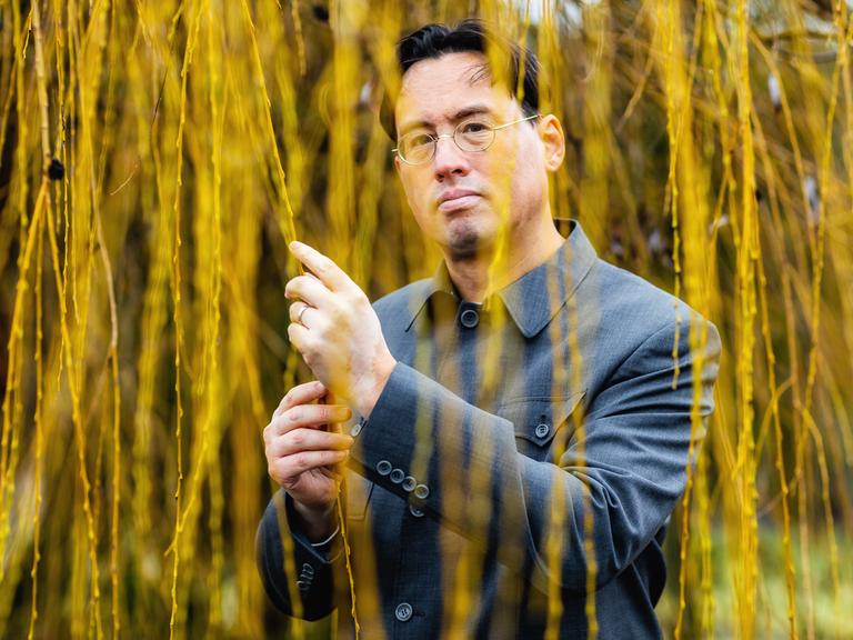 Ein Mann mit chinesisch-amerikanischen Wurzeln steht unter einem Baum und hält herabhängende Weidenzweige in der Hand Ein Mann mit chinesisch-amerikanischen Wurzeln steht unter einem Baum und hält herabhängende Weidenzweige in der Hand