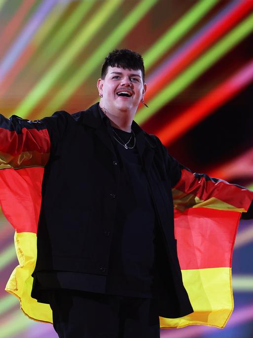 Das Bild zeigt den deutschen Sänger Isaak, der beim Finale des Eurovision Song Contest 2024 die Bühne betritt. Um die Schultern trägt der Sänger eine Deutschlandfahne. Im Hintergrund sind farbige Lichtstrahlen zu sehen.