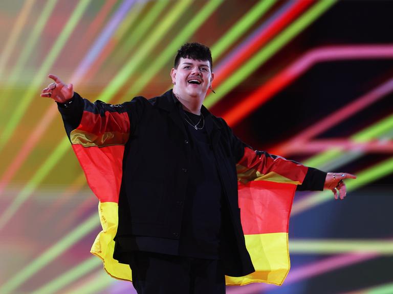 Das Bild zeigt den deutschen Sänger Isaak, der beim Finale des Eurovision Song Contest 2024 die Bühne betritt. Um die Schultern trägt der Sänger eine Deutschlandfahne. Im Hintergrund sind farbige Lichtstrahlen zu sehen.