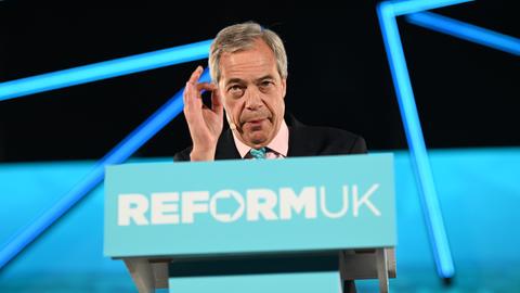 Nigel Farage steht an einem blauen Rednerpult mit der Aufschrift "Reform UK" und spricht ins Publikum.