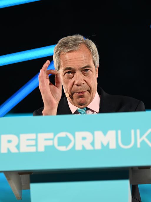Nigel Farage steht an einem blauen Rednerpult mit der Aufschrift "Reform UK" und spricht ins Publikum.