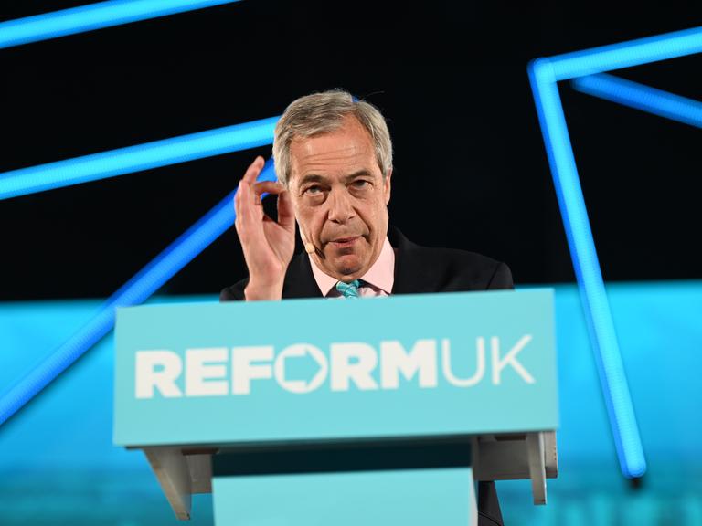 Nigel Farage steht an einem blauen Rednerpult mit der Aufschrift "Reform UK" und spricht ins Publikum.