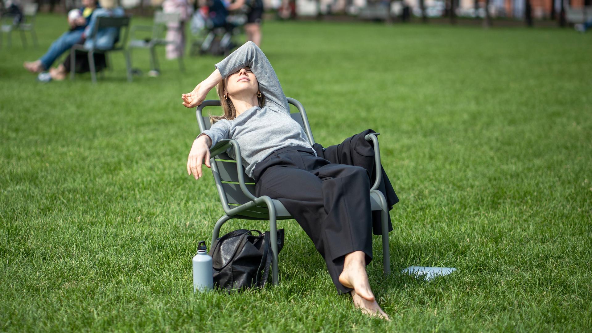 Eine Frau sitzt zurückgelehnt auf einem Stuhl im Park. Mit dem linken Arm verdeckt sie ihr Gesicht. Neben ihr steht eine Trinkflasche im Gras. Sie wirkt erschöpft.