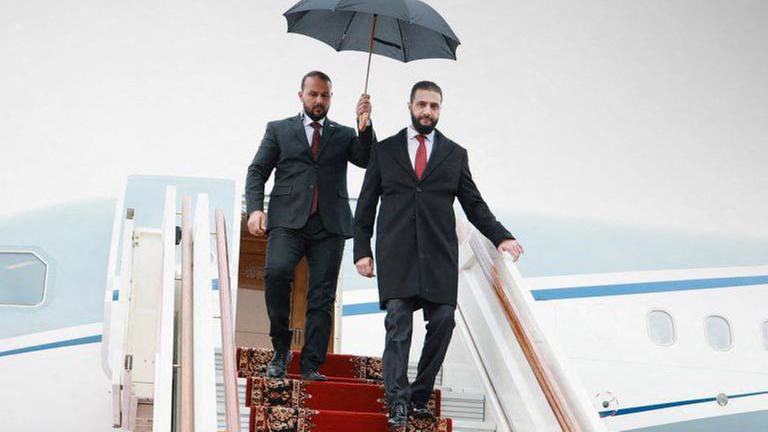 Syriens Übergangspräsident al-Scharaa steigt aus einem Flugzeug aus und geht eine Treppe hinunter. Eine Person hinter ihm hält ihm dabei einen geöffneten Regenschirm über den Kopf.