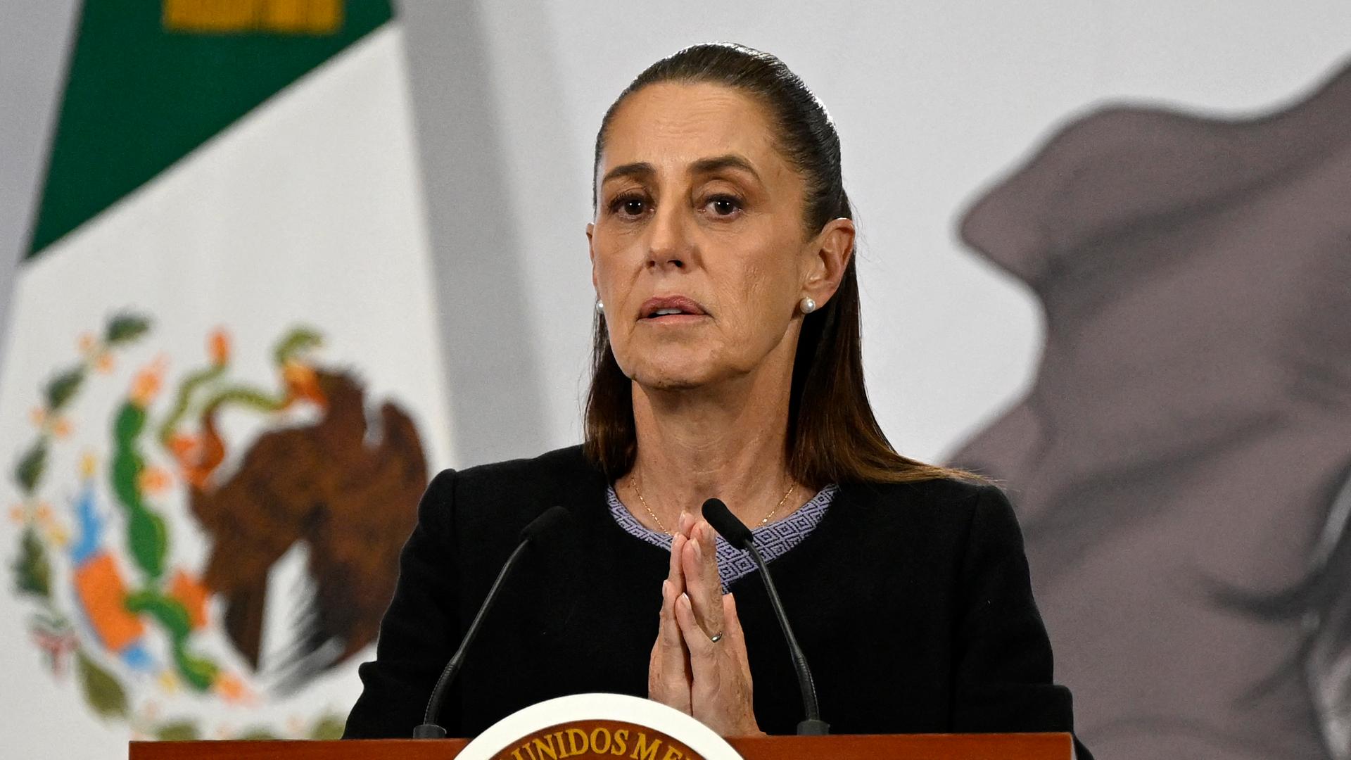 Mexikos Präsidentin Claudia Scheinbaum steht hinter einem Rednerpult und hat die Hände gefaltet.