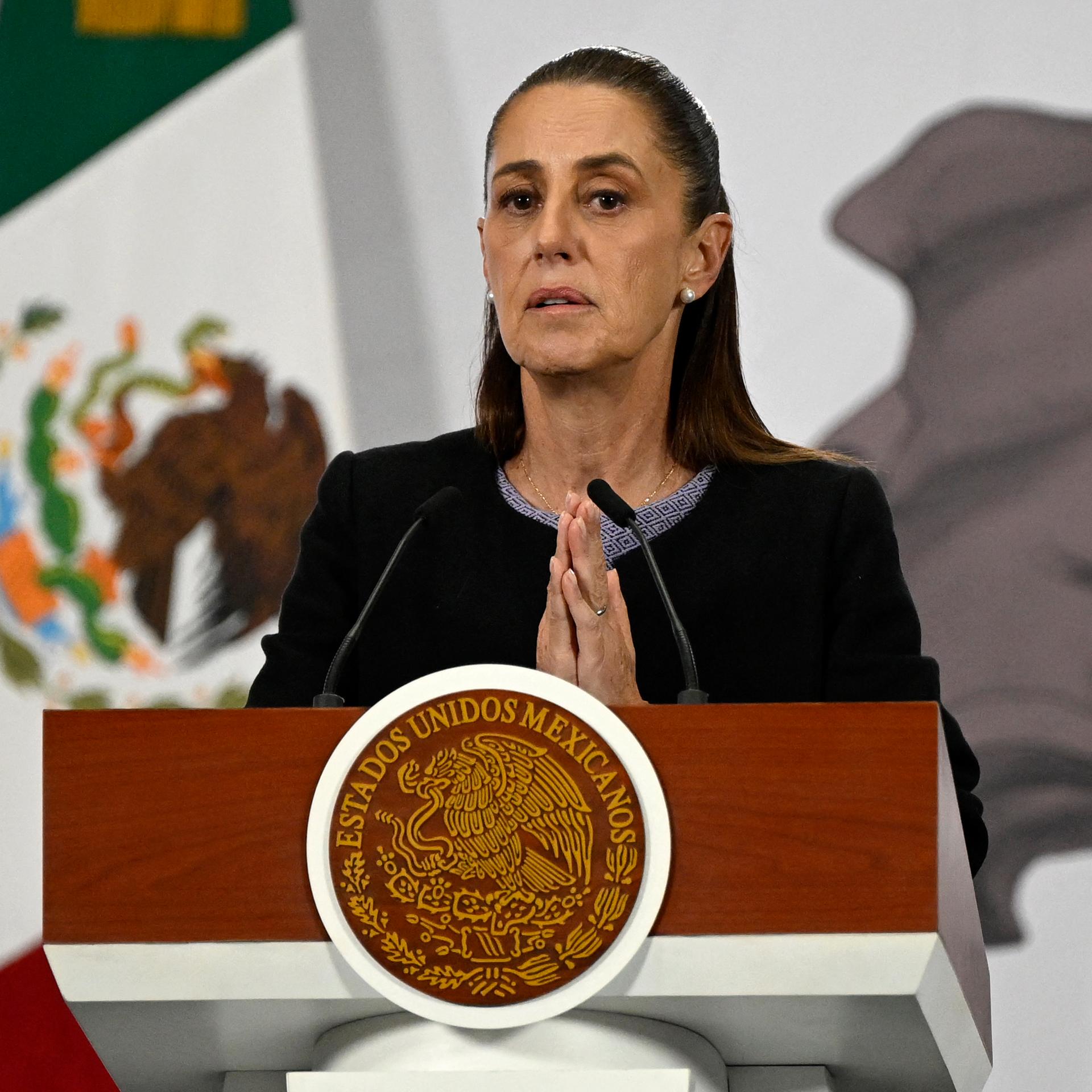Mexikos Präsidentin Claudia Scheinbaum steht hinter einem Rednerpult und hat die Hände gefaltet.
