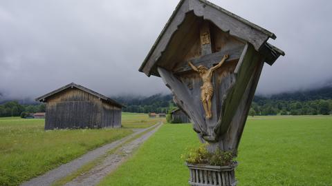 Ein Wegkreuz steht an einem Wanderpfad, im Hintergrund liegt der Wald im Nebel.