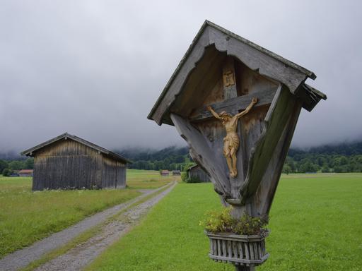 Ein Wegkreuz steht an einem Wanderpfad, im Hintergrund liegt der Wald im Nebel.