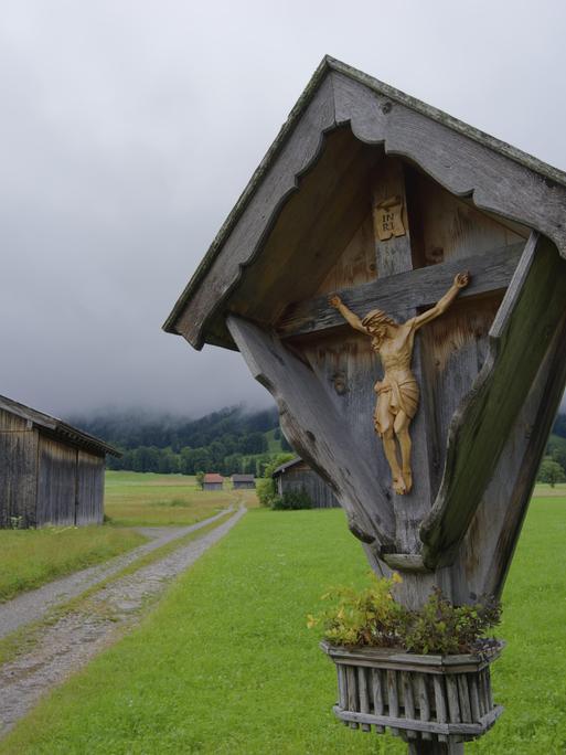 Ein Wegkreuz steht an einem Wanderpfad, im Hintergrund liegt der Wald im Nebel.