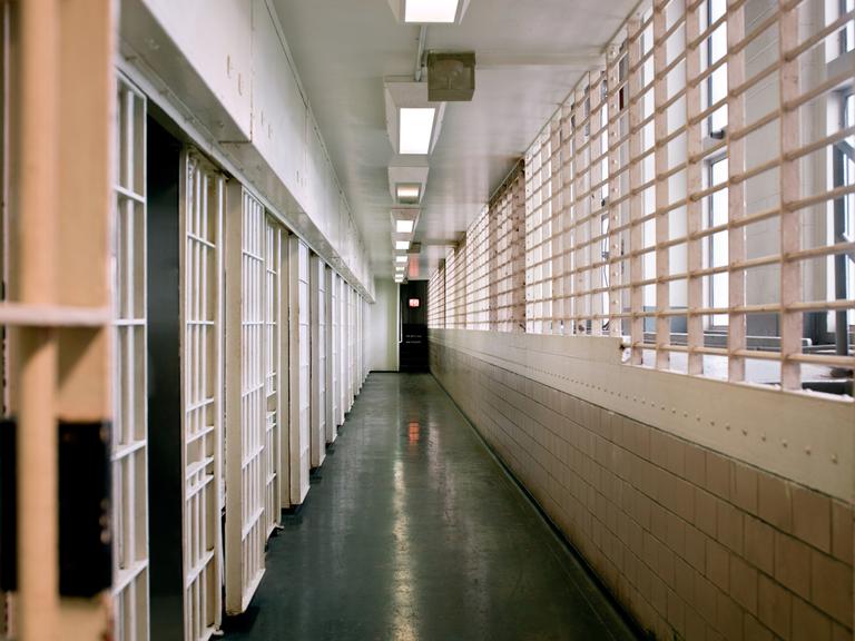 Blick in einen Zellengang im Gefängnis. Die Fenster auf der rechten Seite sind vergittert. Links sind die Zellentüren zu sehen.