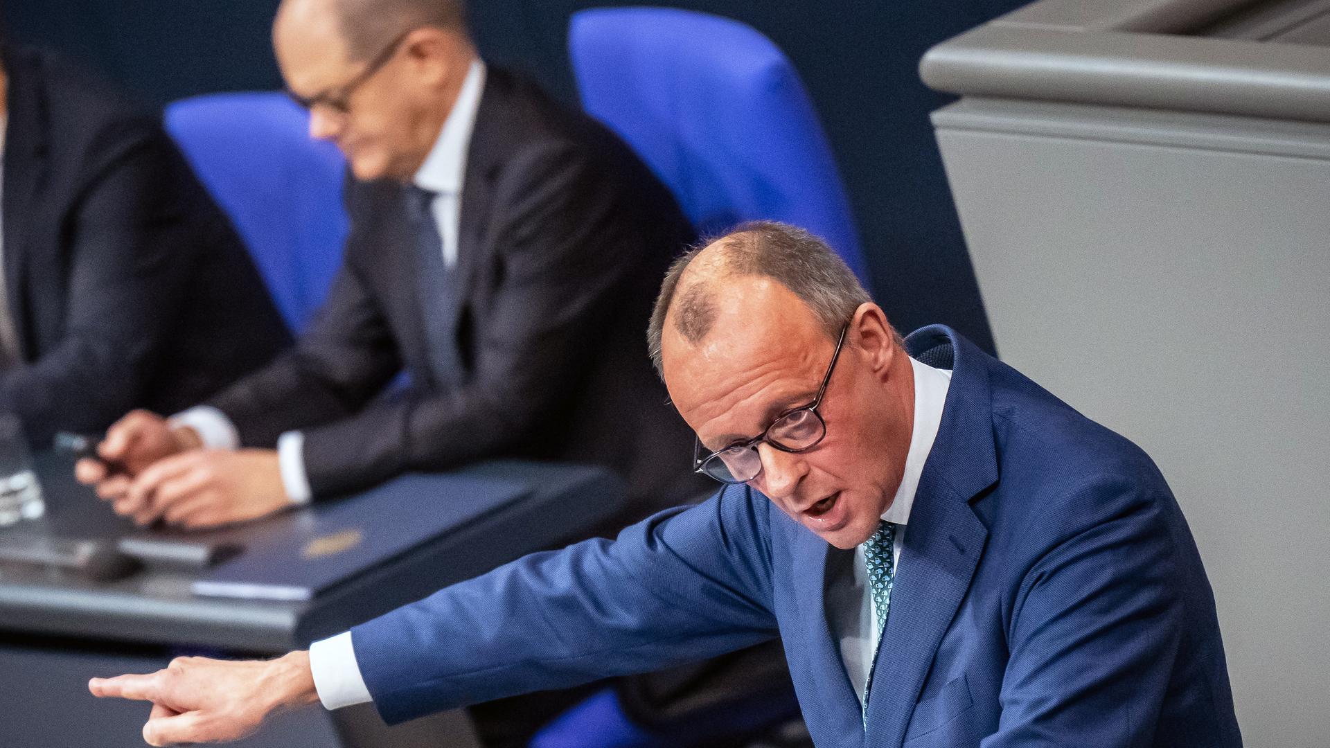 Friedrich Merz spricht im Bundestag. Im Hintergrund schaut Olaf Scholz auf sein Handy.