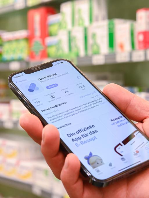 Jemand hält in einer Apotheke ein Smartphone in der Hand, auf dessen Display eine App zur Verarbeitung von E-Rezepten geöffnet ist.
