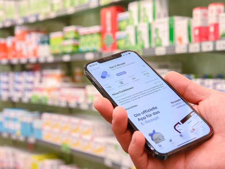 Jemand hält in einer Apotheke ein Smartphone in der Hand, auf dessen Display eine App zur Verarbeitung von E-Rezepten geöffnet ist.