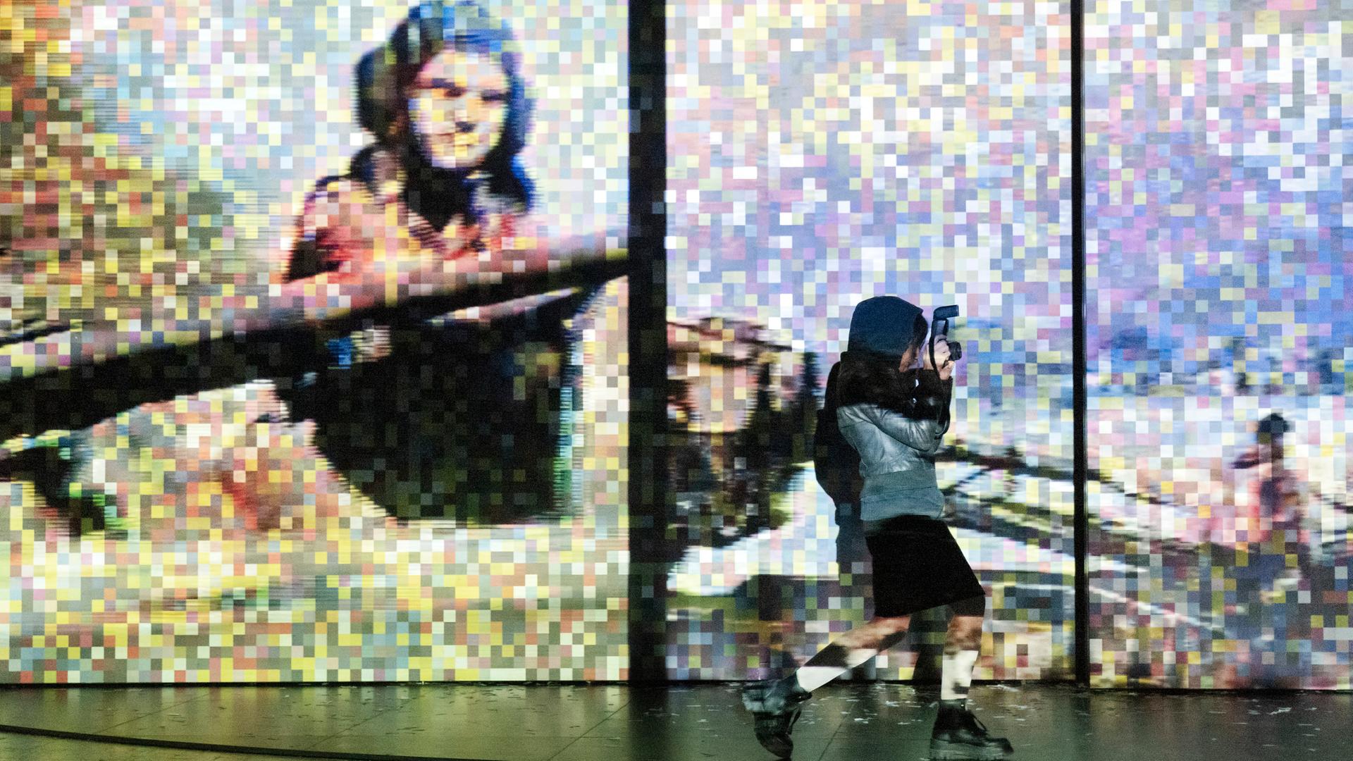 Foto von der Inszenierung: Eine Frau fotografiert im Vordergrund. Im Hintergrund ist ein verpixeltes Bild eines Panzerfahres in Aktion zu sehen.