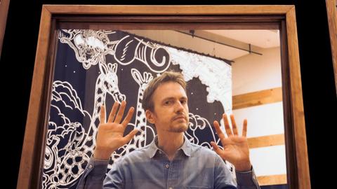 Ein schmaler Mann steht in einem quadratischen Bilderrahmen und hält die Hände erhoben und zeigt seine Finger. Sein Blick ist etwas verschmitzt.