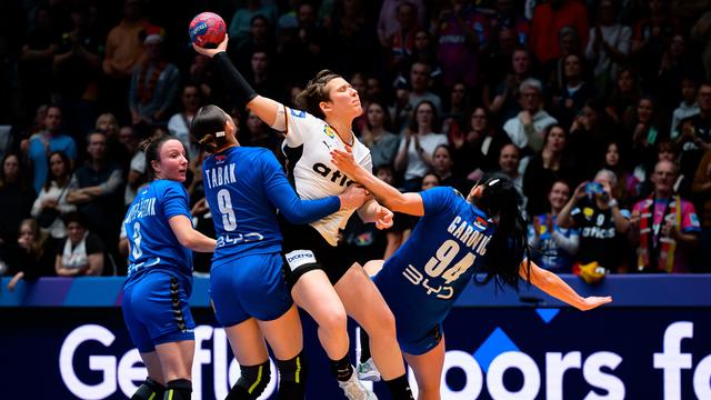 Die deutsche Handball-Spielerin Alina Grijseels wirft den Ball, obwohl sie von zwei Spielerinnen der serbischen Mannschaft heftig festgehalten wird.