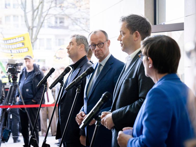 Markus Söder, Friedrich Merz, Lars Klingbeil und Saskia Esken stehen bei einer Pressekonferenz vor den Mikrofonen. Friedrich Merz schaut seitlich in die Kamera. 