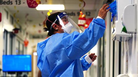 Eine im medizinischen Bereich arbeitende Person steht in einem Krankenhausflur in kompletter Covid-Schutzmontur und entnimmt einem Handschuhspender blaue Gummihandschuhe.