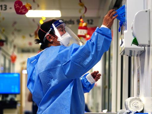 Eine im medizinischen Bereich arbeitende Person steht in einem Krankenhausflur in kompletter Covid-Schutzmontur und entnimmt einem Handschuhspender blaue Gummihandschuhe.
