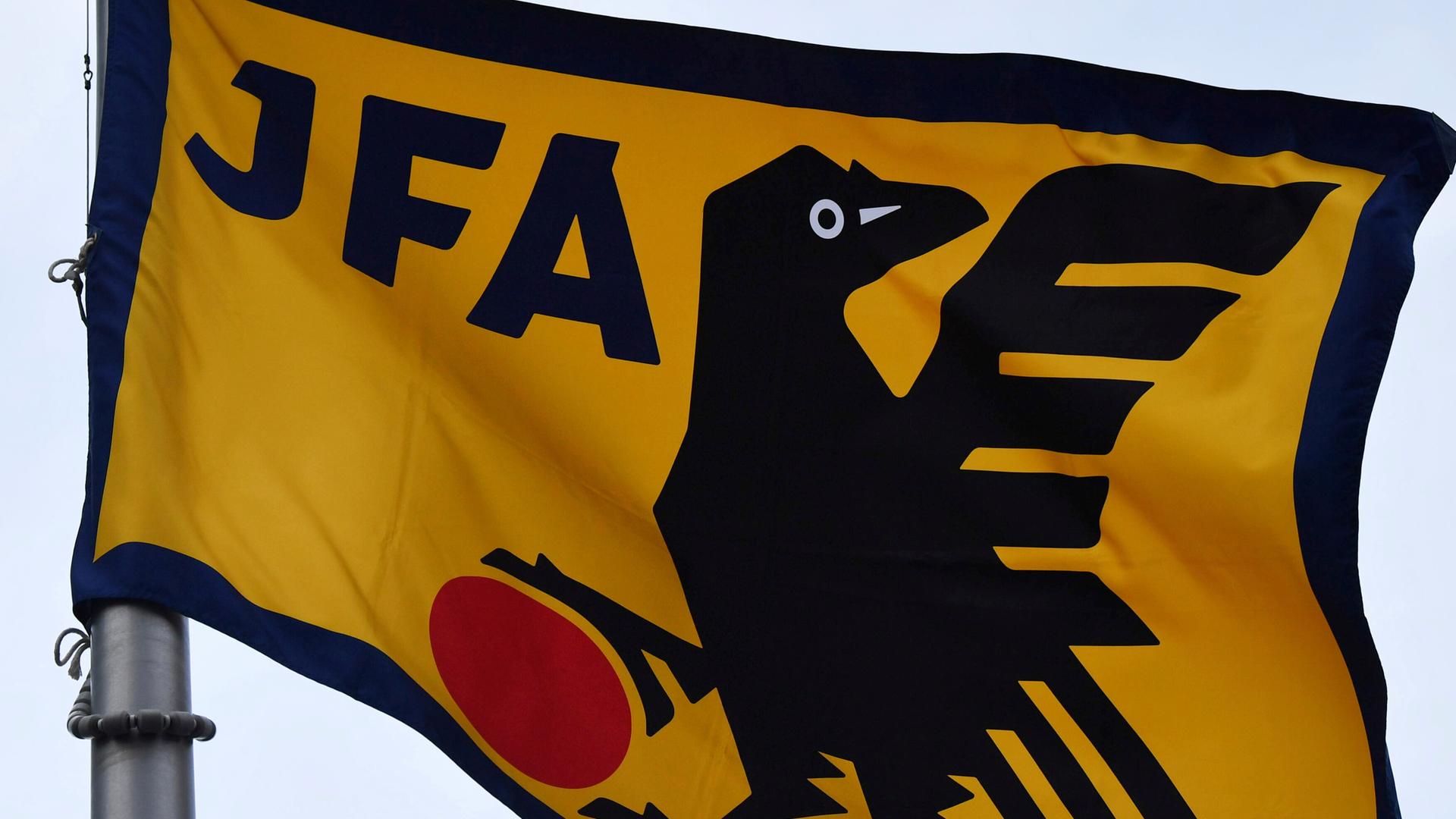 Auf einer gelben Flagge mit schwarzem Rand steht in schwerzer Schrift "JFA". Daneben eine schwarze stilisierte Krähe, die eine Kralle auf einem roten Ball hat. Auf einer gelben Flagge mit schwarzem Rand steht in schwerzer Schrift "JFA". Daneben eine schwarze stilisierte Krähe, die eine Kralle auf einem roten Ball hat.