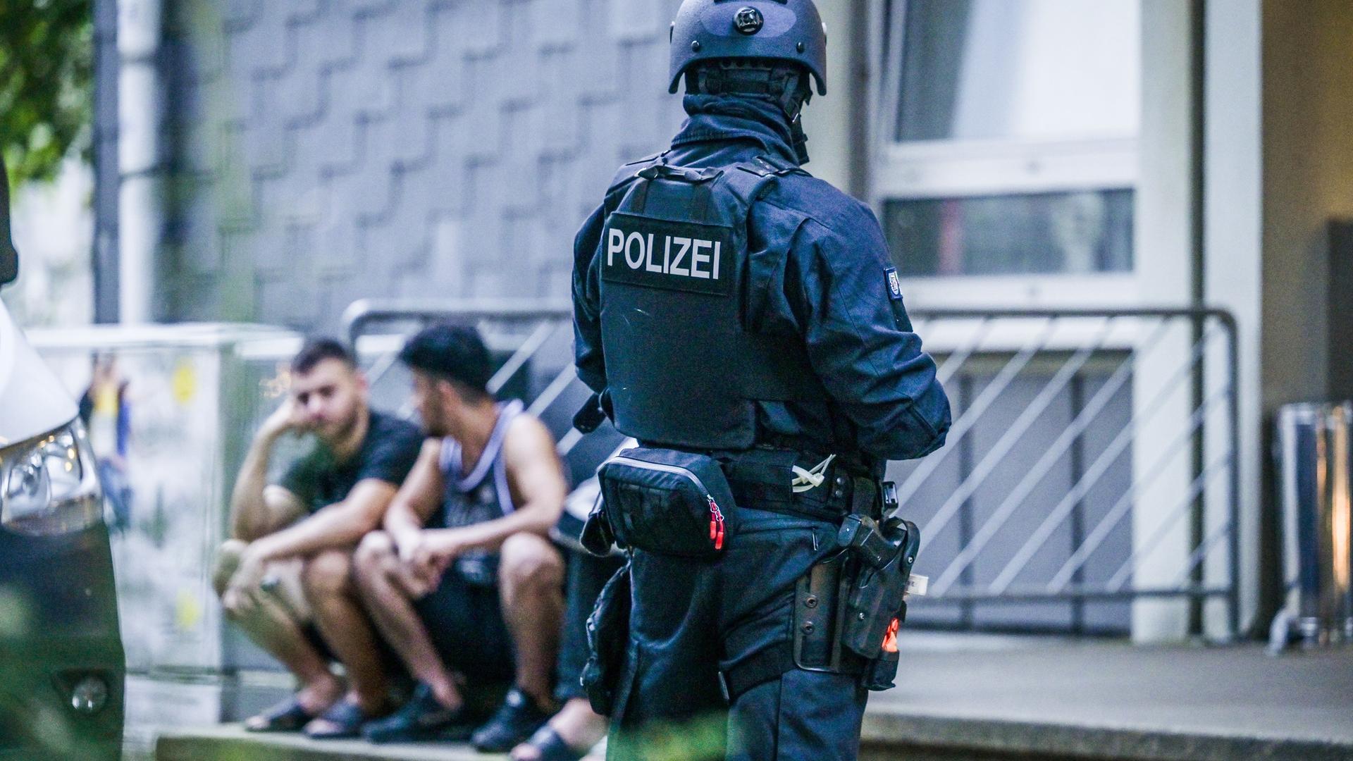 Polizisten in Sturmausrüstung stehen vor einem Gebäude, auf dessen Treppenstufen einige Männer in kurzer Sommerkleidung sitzen.