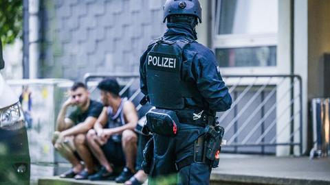 Polizisten in Sturmausrüstung stehen vor einem Gebäude, auf dessen Treppenstufen einige Männer in kurzer Sommerkleidung sitzen.