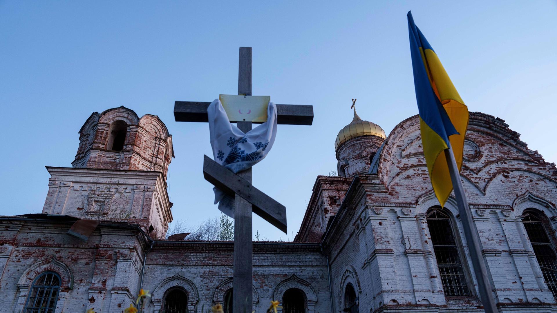 Ein orthodoxes Kreuz steht vor einer zerstörten Kirche in dem ukrainischen Dorf Lukaschiwka. Rechts daneben hängt eine ukrainische Flagge. Ein orthodoxes Kreuz steht vor einer zerstörten Kirche in dem ukrainischen Dorf Lukaschiwka. Rechts daneben hängt eine ukrainische Flagge.