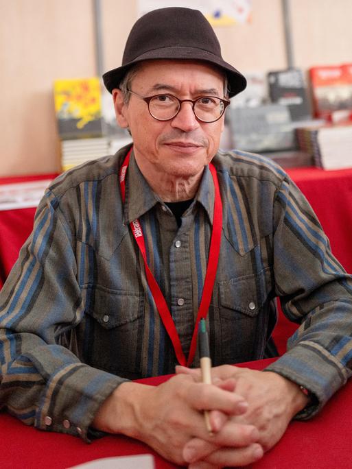 Joe Sacco posiert beim Festival America in Vincennes, Frankreich, mit Hut an einem Tisch sitzend für ein Foto. Joe Sacco posiert beim Festival America in Vincennes, Frankreich, mit Hut an einem Tisch sitzend für ein Foto.