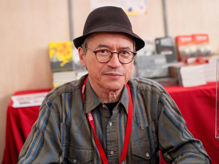 Joe Sacco posiert beim Festival America in Vincennes, Frankreich, mit Hut an einem Tisch sitzend für ein Foto. Joe Sacco posiert beim Festival America in Vincennes, Frankreich, mit Hut an einem Tisch sitzend für ein Foto.