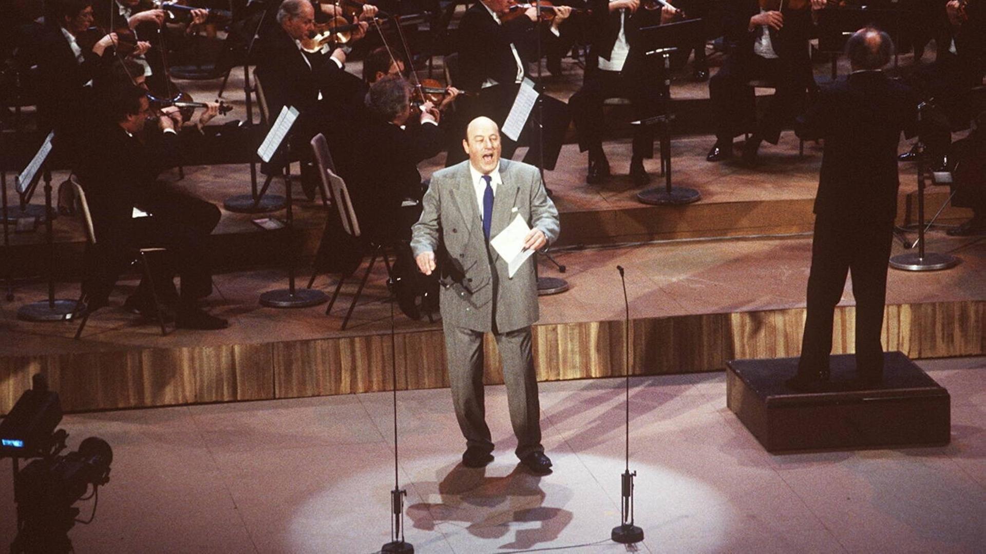 Manfred Krug steht bei einer Preisverleihung im grauen Anzug singend auf der Bühne, ein Orchester hinter sich. Manfred Krug steht bei einer Preisverleihung im grauen Anzug singend auf der Bühne, ein Orchester hinter sich.