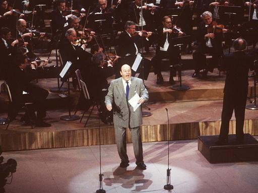 Manfred Krug steht bei einer Preisverleihung im grauen Anzug singend auf der Bühne, ein Orchester hinter sich.