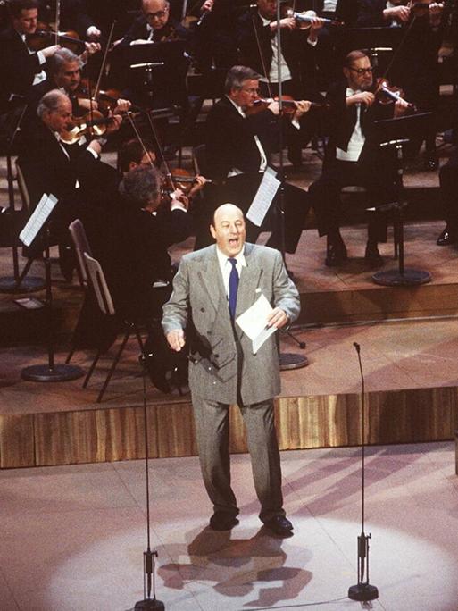 Manfred Krug steht bei einer Preisverleihung im grauen Anzug singend auf der Bühne, ein Orchester hinter sich.