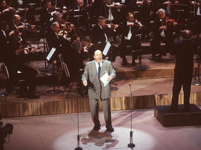 Manfred Krug steht bei einer Preisverleihung im grauen Anzug singend auf der Bühne, ein Orchester hinter sich.
