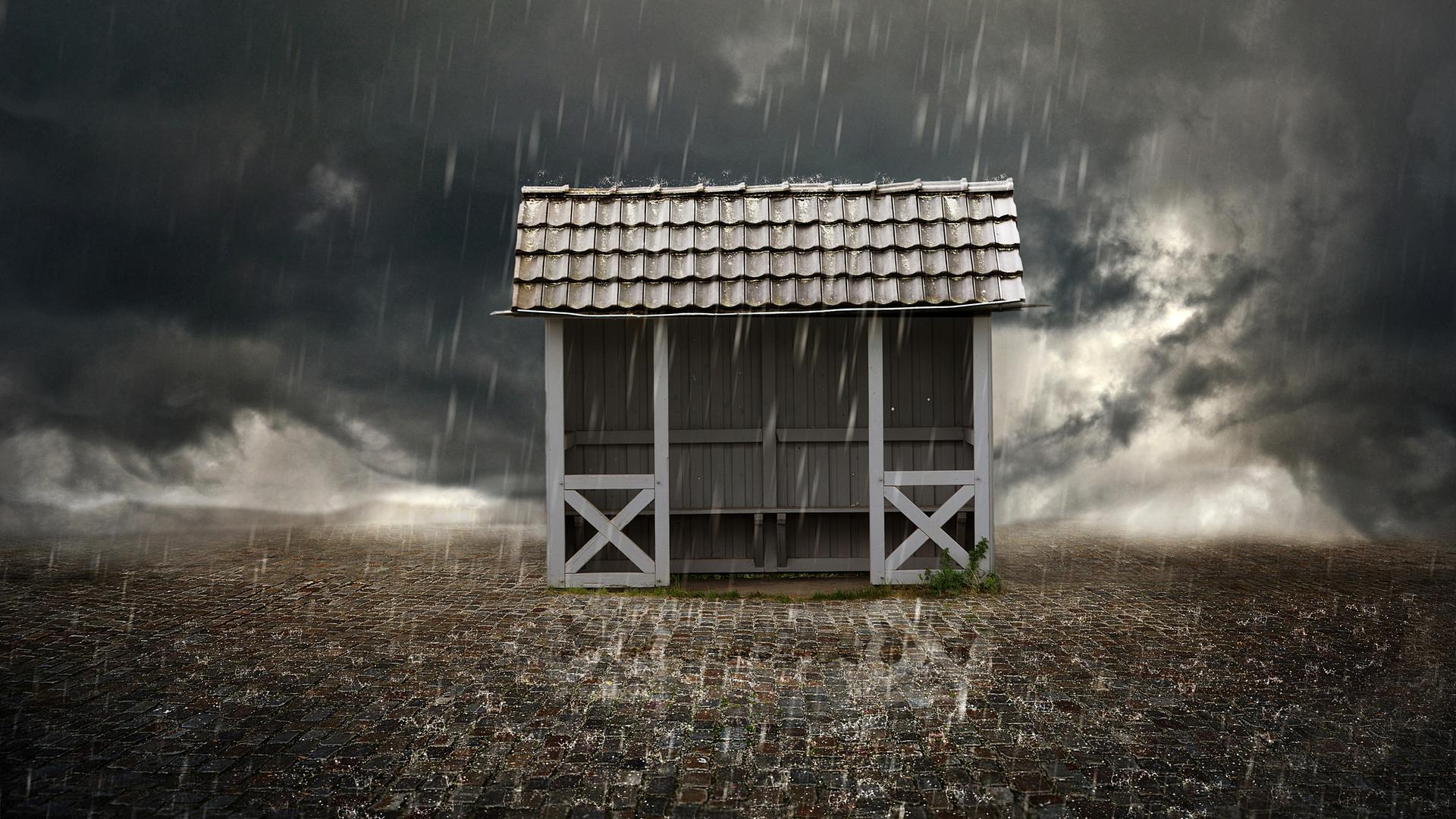 Ein Wartehäuschen einer Bushaltestelle steht bei schwerem Gewitter im Starkregen. Auf dem Boden im Vordergrund sind die prasselnden Regentropfen zu sehen.
