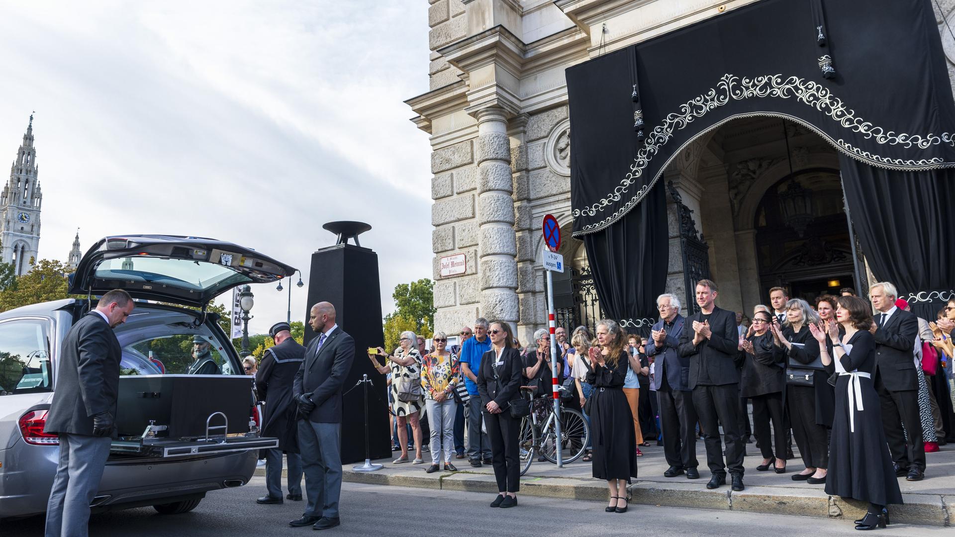 Der Sarg mit dem Leichnam wurde extra für die Trauerfeier von Berlin nach Wien gebracht. Ein Leichenwagen steht vor dem Burgtheater; der Eingang ist mit einem schwarzen Vorhang abgehängt; der Sarg ist im Auto zu sehen; viele Menschen stehen dort.
Der Sarg mit dem Leichnam wurde extra für die Trauerfeier von Berlin nach Wien gebracht. Ein Leichenwagen steht vor dem Burgtheater; der Eingang ist mit einem schwarzen Vorhang abgehängt; der Sarg ist im Auto zu sehen; viele Menschen stehen dort.