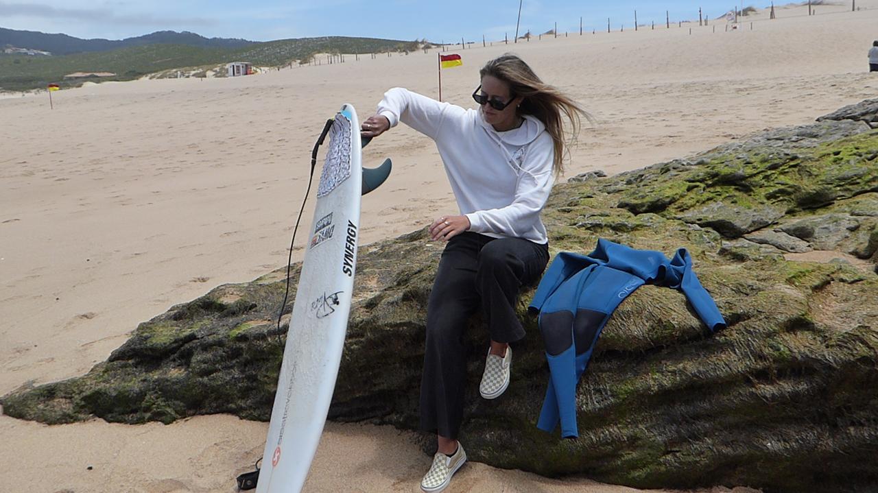 Zu sehen ist Camilla Kemp, sie sitzt auf einem Felsen am Strand, neben ihr liegt ihr Neopren-Anzug, vor sich hält sie ihr weißes Shortboard.