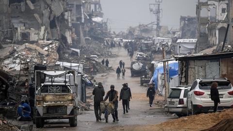 Palästinenser gehen im Regen eine Straße mit kriegszerstörten Gebäuden in Gaza-Stadt entlang.