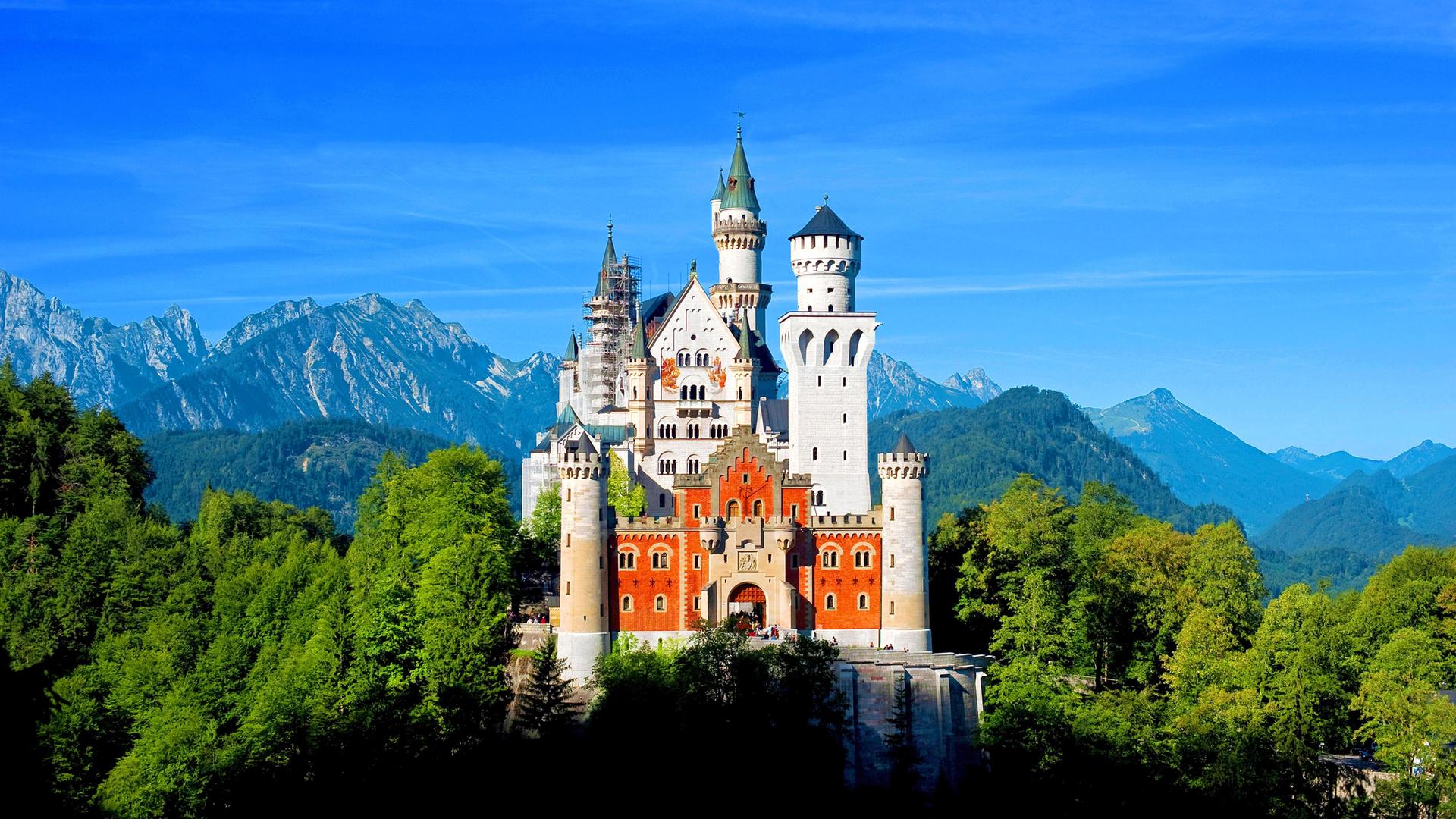Das Schloss Neuschwanstein mit seinen Türmen liegt im Sonnenschein. Im Hintergrund sind Berge, Wald uns blauer Himmel zu sehen. Das Schloss Neuschwanstein mit seinen Türmen liegt im Sonnenschein. Im Hintergrund sind Berge, Wald uns blauer Himmel zu sehen.