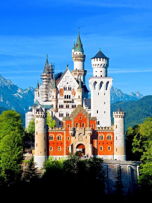 Das Schloss Neuschwanstein mit seinen Türmen liegt im Sonnenschein. Im Hintergrund sind Berge, Wald und blauer Himmel zu sehen.